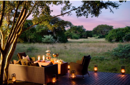 hurricane lamps light up the deck of a safari lodge where afternoon tea is set on the table over looking the bush veld.