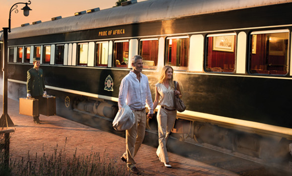 A couple walk along the platform ready to board the luxury train with a porter behind carrying their suitcases.