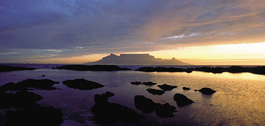 Table Mountain as sun set with yellow sky and silhouette of rocks in the sea.