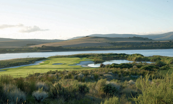 Lagoon surrounding the green and fairways of a golf course with hills beyond.