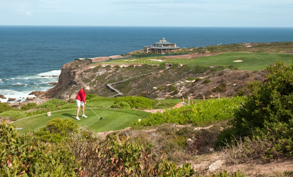 Lady in red top teeing off from 18th tee box across the fynbos towards the clubhouse with ocean in the distance.