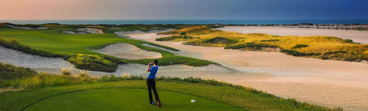 Golfer hits a tee shot past the white sand bunkers towards the ocean.