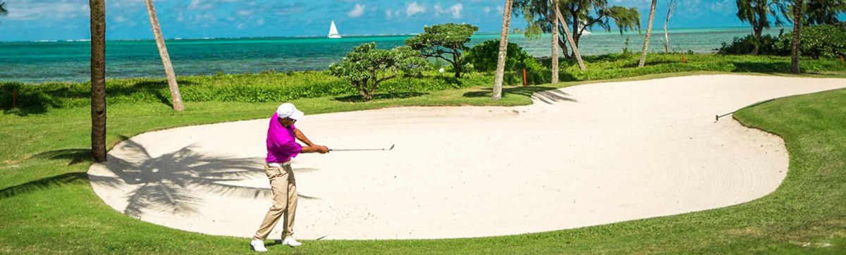 Golfer hitting his ball from the sand bunker as a yacht sails past on the azure blue sea in Mauritius