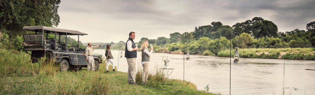 Couple drinking champagne and looking at a river as part of a safari game drive.