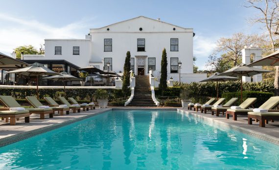 swimming pool lined with sun loungers and umbrellas with manor house in the back ground.